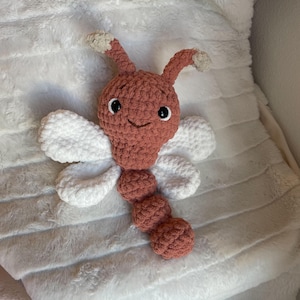 May include: A crocheted plush toy shaped like a dragonfly with a dusty rose-colored head and segmented body, two white wings on each side, and two antennae with beige tips. The toy features black safety eyes and a stitched smile.