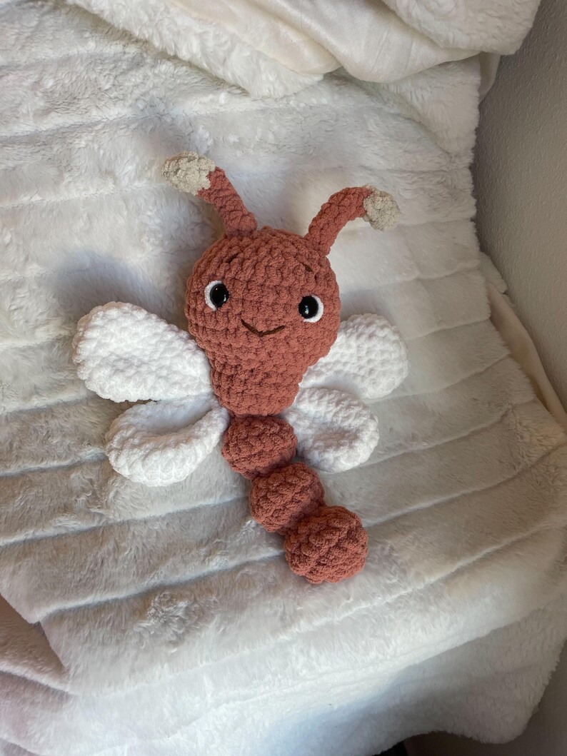 May include: A crocheted plush toy shaped like a dragonfly with a dusty rose-colored head and segmented body, two white wings on each side, and two antennae with beige tips. The toy features black safety eyes and a stitched smile.