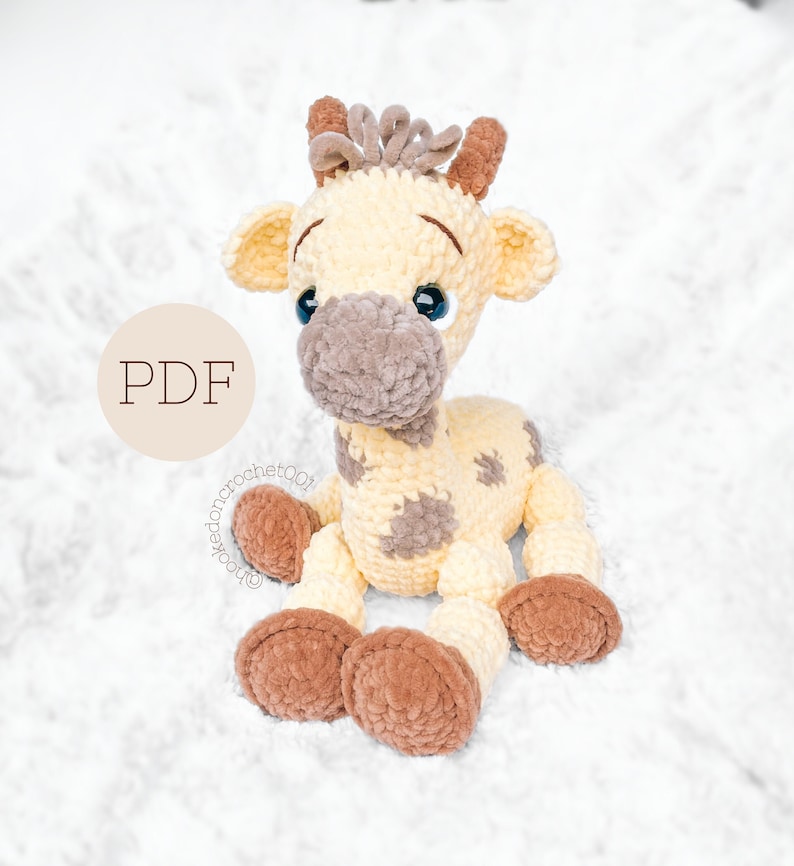 May include: A crocheted giraffe amigurumi toy with brown spots, brown hooves, and blue eyes. The giraffe is sitting on a white fluffy surface. The text "PDF" is visible in the image.