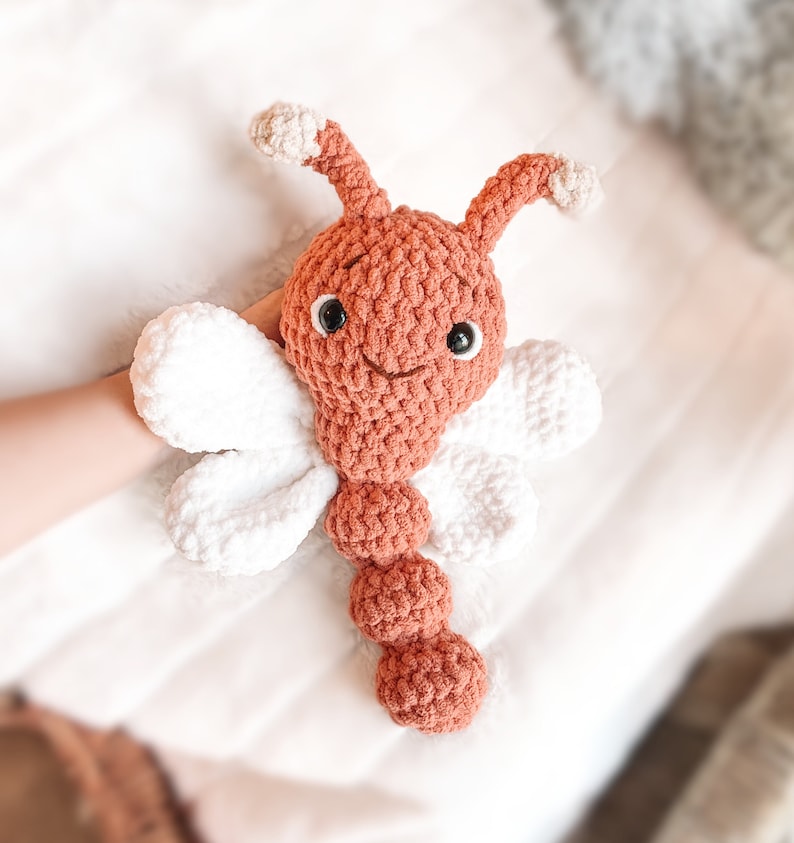 May include: A handmade crochet plush toy shaped like a butterfly with a terracotta-colored head and segmented body, white wings, and two antennae with white tips. The toy features black plastic safety eyes and a stitched smile.