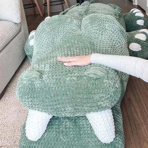 May include: A large, plush, green alligator-shaped pillow. The pillow has white teeth and white accents on the feet. The alligator is lying on a wooden floor. A person's hand is resting on the pillow.
