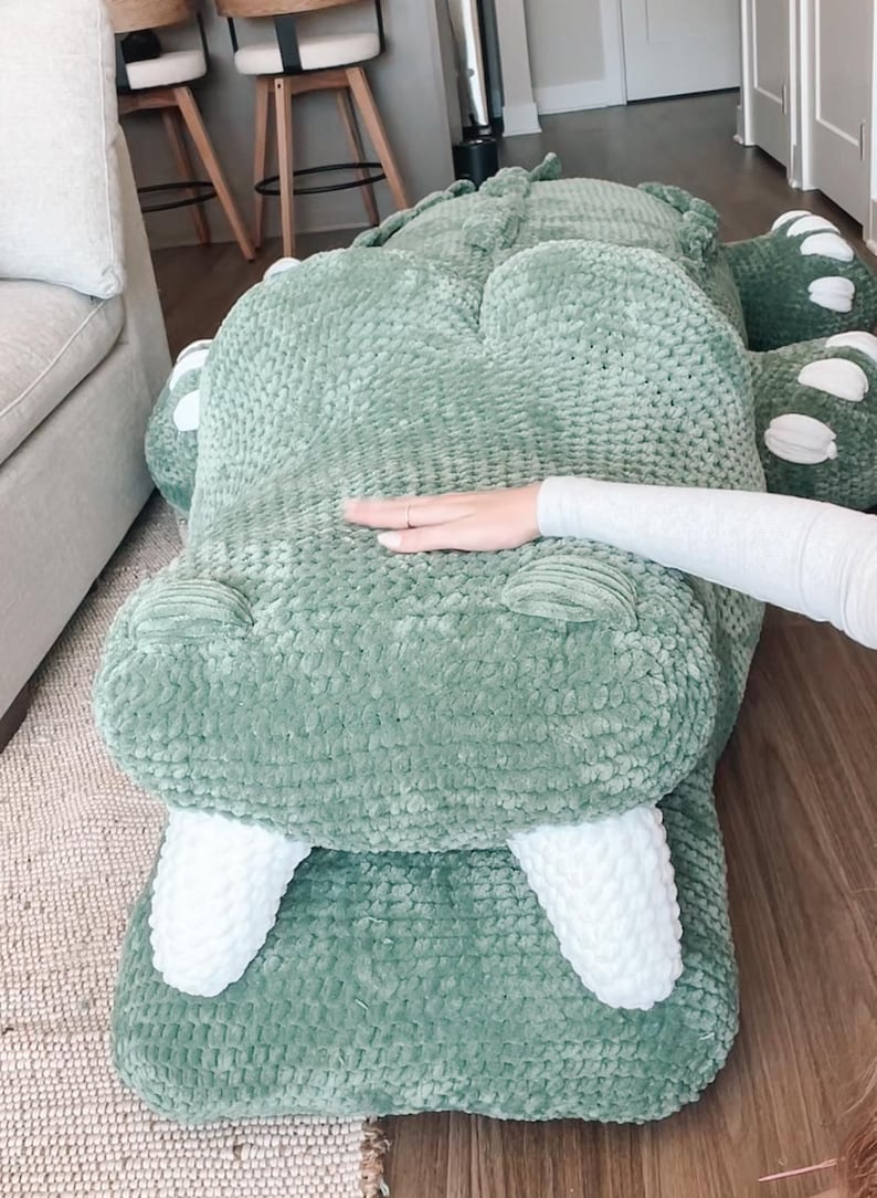 May include: A large, plush, green alligator-shaped pillow. The pillow has white teeth and white accents on the feet. The alligator is lying on a wooden floor. A person's hand is resting on the pillow.