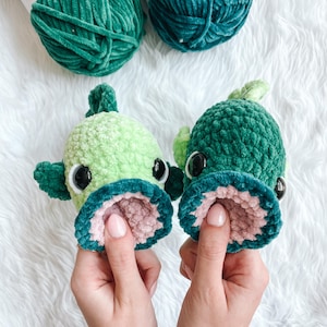 Op de afbeelding: Twee handgemaakte gehaakte visjes in groentinten, met roze en blauwe accenten. De vissen hebben grote, zwarte ogen en open monden. Op de achtergrond liggen twee bollen groen garen.