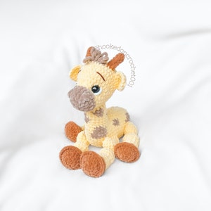 May include: A crocheted giraffe toy with brown and yellow yarn. The giraffe has brown spots and a brown nose. The giraffe is sitting on a white surface.