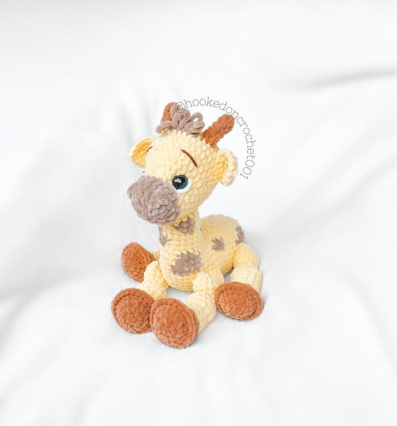 May include: A crocheted giraffe toy with brown and yellow yarn. The giraffe has brown spots and a brown nose. The giraffe is sitting on a white surface.