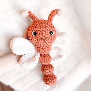 May include: A handmade crochet dragonfly plush toy featuring a rust-colored head and segmented body, two white wings on each side, and two rust-colored antennae with light beige tips. The toy has black plastic safety eyes and a stitched mouth.