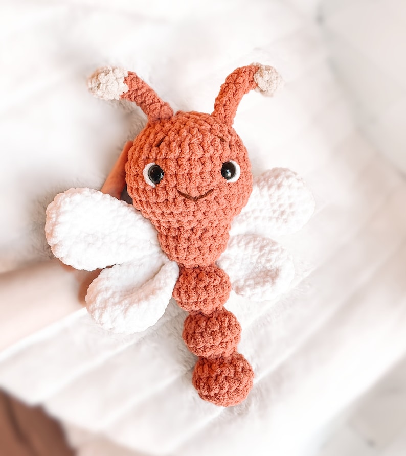May include: A handmade crochet dragonfly plush toy featuring a rust-colored head and segmented body, two white wings on each side, and two rust-colored antennae with light beige tips. The toy has black plastic safety eyes and a stitched mouth.