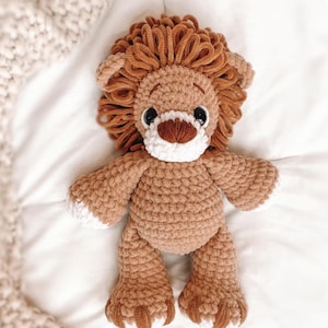 May include: A handmade crochet lion stuffed animal. The lion has a brown body, a fluffy brown mane, and white accents on its face and paws. It has black eyes and a brown nose. The toy is made of soft yarn and is designed to be cuddly.