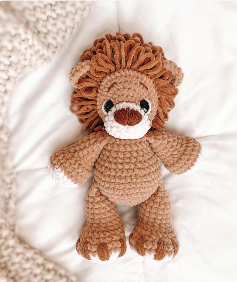 May include: A handmade crochet lion stuffed animal. The lion has a brown body, a fluffy brown mane, and white accents on its face and paws. It has black eyes and a brown nose. The toy is made of soft yarn and is designed to be cuddly.