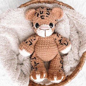 May include: A handmade crochet tiger plush toy in a woven basket. The plush is tan with black markings, white accents on the paws and muzzle, and black eyes. The toy is sitting on a fluffy, light-colored blanket.