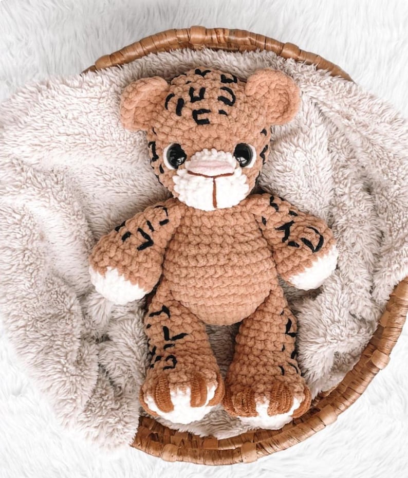 May include: A handmade crochet tiger plush toy in a woven basket. The plush is tan with black markings, white accents on the paws and muzzle, and black eyes. The toy is sitting on a fluffy, light-colored blanket.