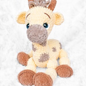 May include: A plush giraffe toy in cream, brown, and tan. The handmade stuffed animal has a soft, textured appearance with brown spots, large blue eyes, and brown hooves. The giraffe is sitting on a white, fluffy surface.
