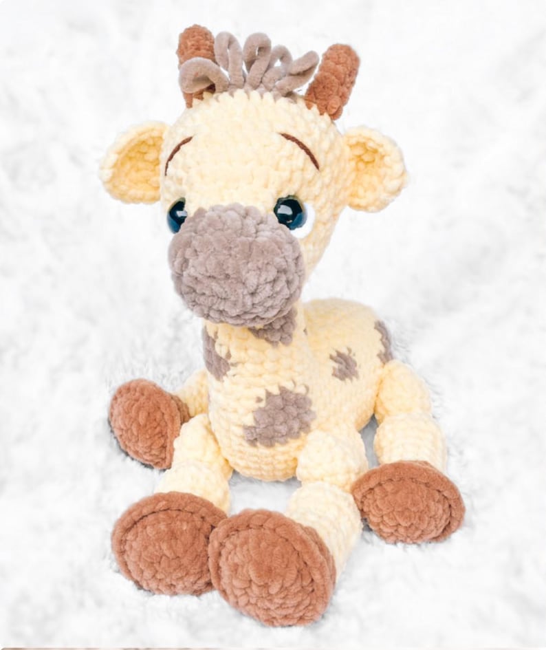 May include: A plush giraffe toy in cream, brown, and tan. The handmade stuffed animal has a soft, textured appearance with brown spots, large blue eyes, and brown hooves. The giraffe is sitting on a white, fluffy surface.
