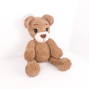 Peut inclure: Un ours en peluche crochet&eacute; marron avec des accents de fil blanc pour le nez et les yeux. L'ours est assis les jambes tendues.