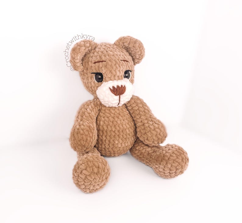 Peut inclure: Un ours en peluche crochet&eacute; marron avec des accents de fil blanc pour le nez et les yeux. L'ours est assis les jambes tendues.