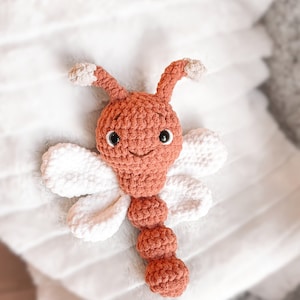 May include: A crocheted stuffed toy shaped like a dragonfly with a rust-colored head, body, and antennae, featuring white crocheted wings. The toy has two black plastic safety eyes and a stitched mouth on its round head.