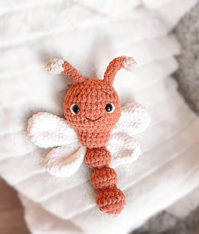 May include: A crocheted stuffed toy shaped like a dragonfly with a rust-colored head, body, and antennae, featuring white crocheted wings. The toy has two black plastic safety eyes and a stitched mouth on its round head.