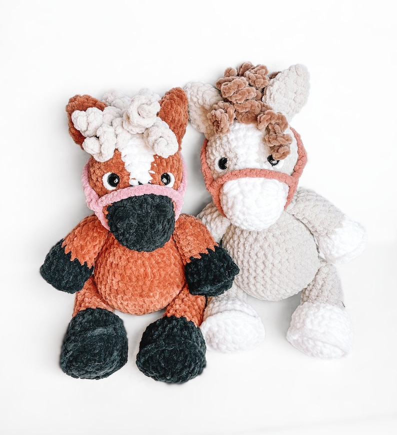 May include: Two crocheted plush horse toys. One is brown with black hooves and a pink nose, the other is white with brown fur and a pink nose.