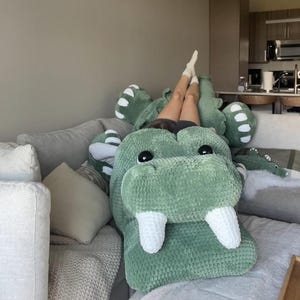 May include: A large, green plush alligator-shaped pillow with white teeth and black eyes rests on a gray sofa. The pillow is made of a soft, textured material. The alligator pillow is a unique home decor item.