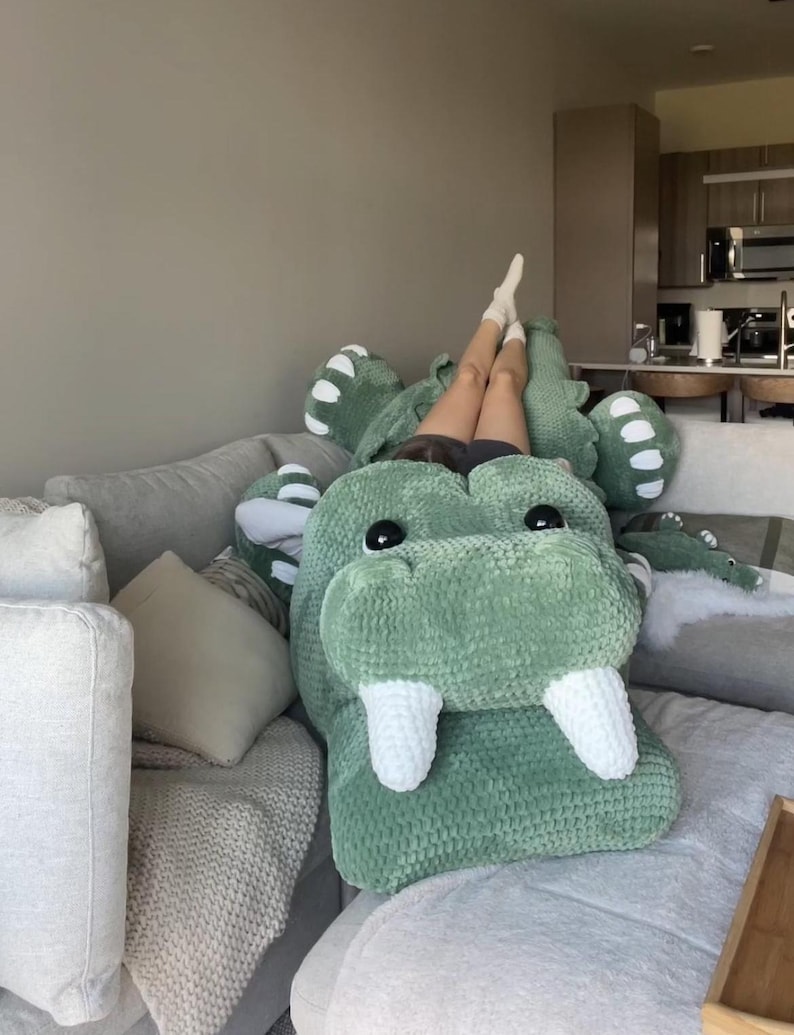 May include: A large, green plush alligator-shaped pillow with white teeth and black eyes rests on a gray sofa. The pillow is made of a soft, textured material. The alligator pillow is a unique home decor item.