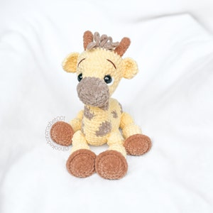 May include: A crocheted giraffe plush toy with brown spots and brown hooves. The giraffe is sitting on a white surface.
