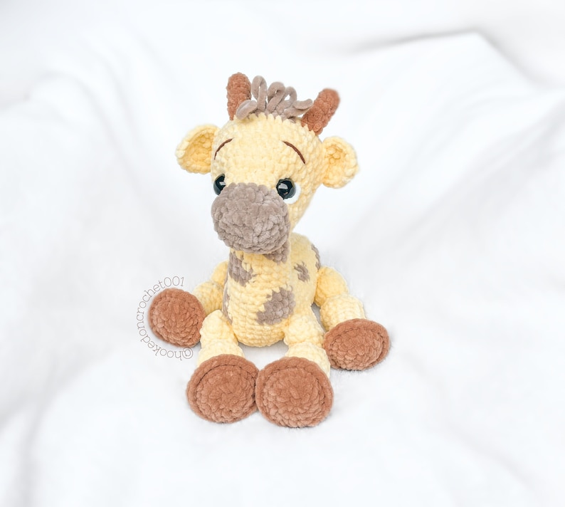 May include: A crocheted giraffe plush toy with brown spots and brown hooves. The giraffe is sitting on a white surface.