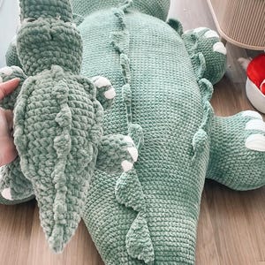 May include: A large, soft, green crocheted alligator-shaped pillow and a smaller matching alligator plush toy. The larger pillow has a detailed spine and white accents on the feet. The plush toy is held in a hand.