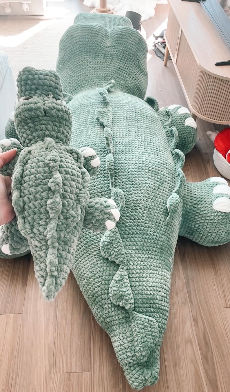 May include: A large, soft, green crocheted alligator-shaped pillow and a smaller matching alligator plush toy. The larger pillow has a detailed spine and white accents on the feet. The plush toy is held in a hand.