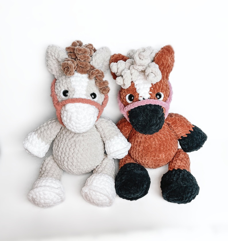 May include: Two crocheted stuffed horses, one gray and one brown, with pink and black details. The horses have fluffy manes and tails.