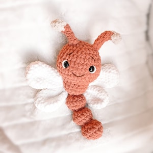 May include: A crocheted stuffed toy shaped like a dragonfly with a rust-colored head, body, and antennae, featuring white wings and two black plastic eyes. The antennae tips are light beige, and the body consists of three rounded segments.