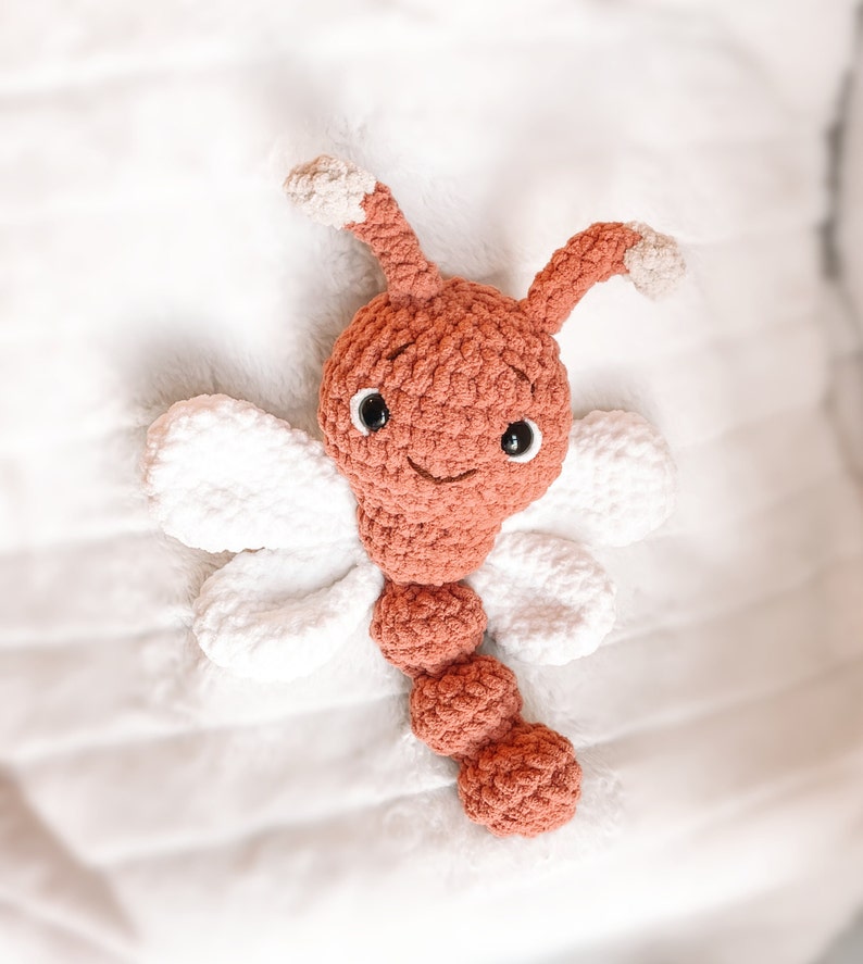 May include: A crocheted stuffed toy shaped like a dragonfly with a rust-colored head, body, and antennae, featuring white wings and two black plastic eyes. The antennae tips are light beige, and the body consists of three rounded segments.