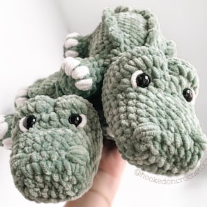 May include: Crocheted green dinosaur toy with white accents on the feet and belly. The dinosaur has black safety eyes and is made of a soft, textured yarn. The toy is handmade and suitable for children.