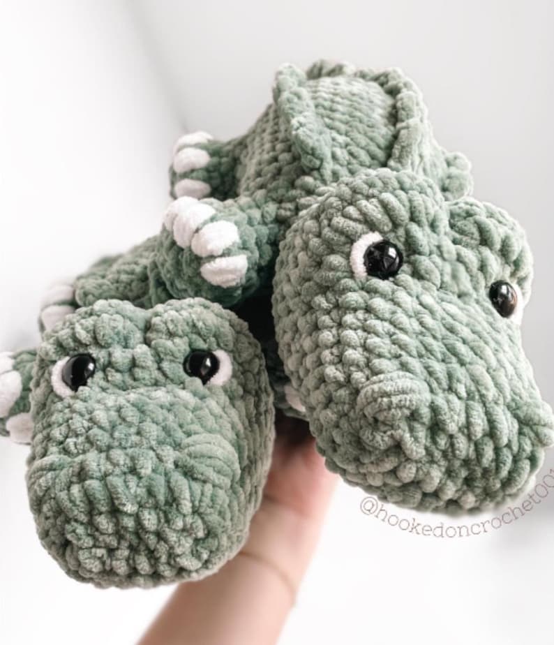 May include: Crocheted green dinosaur toy with white accents on the feet and belly. The dinosaur has black safety eyes and is made of a soft, textured yarn. The toy is handmade and suitable for children.