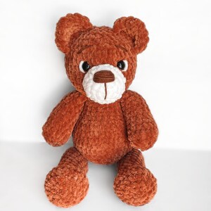 Peut inclure: Un ours en peluche crochet&eacute; marron avec des traits du visage blancs et des yeux noirs. L'ours est assis les jambes crois&eacute;es.