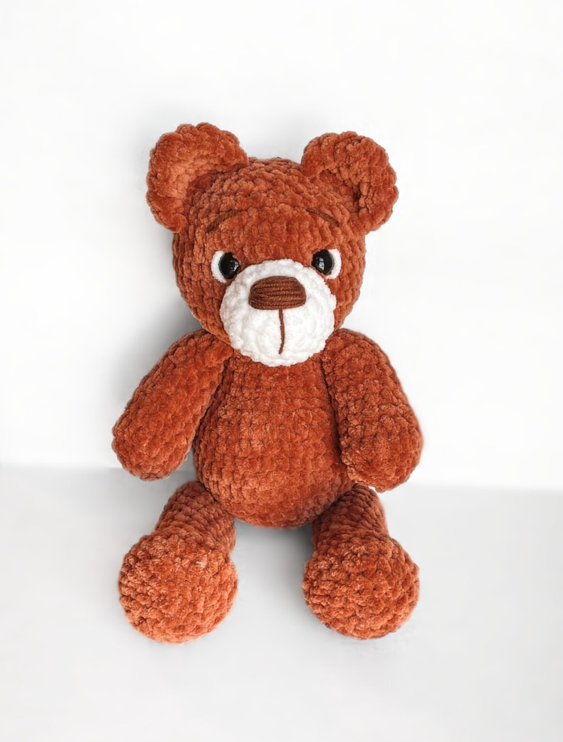 Peut inclure: Un ours en peluche crochet&eacute; marron avec des traits du visage blancs et des yeux noirs. L'ours est assis les jambes crois&eacute;es.
