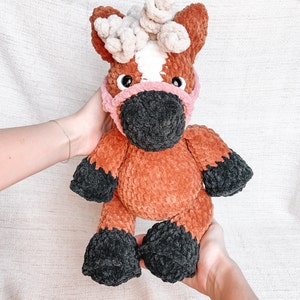 May include: A brown and white crocheted horse toy with a pink and black face mask. The horse has a white star on its forehead and black hooves.