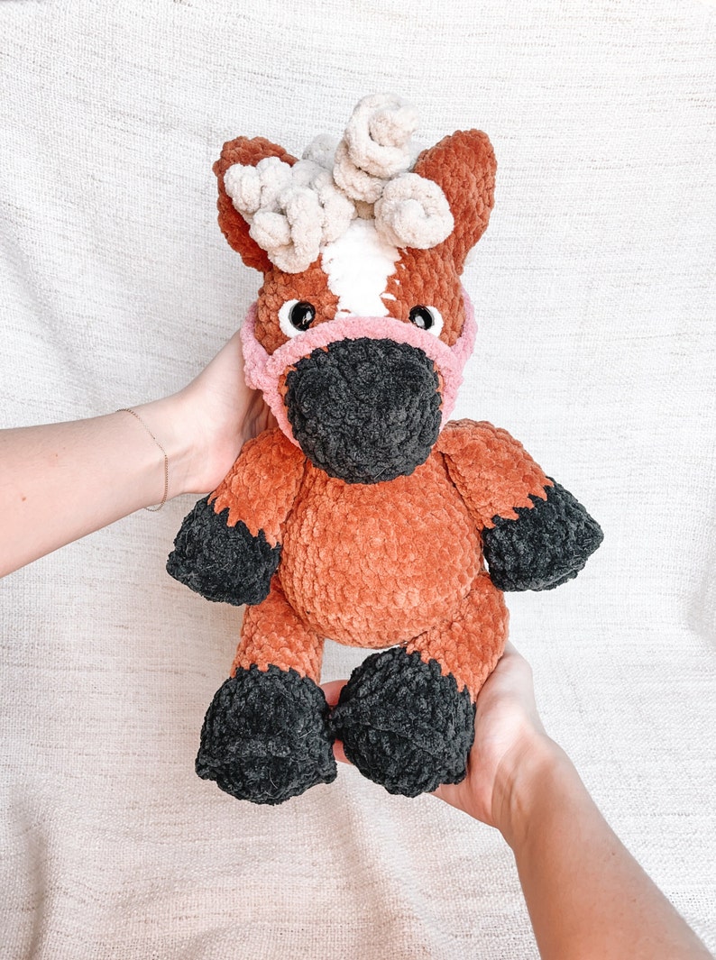 May include: A brown and white crocheted horse toy with a pink and black face mask. The horse has a white star on its forehead and black hooves.