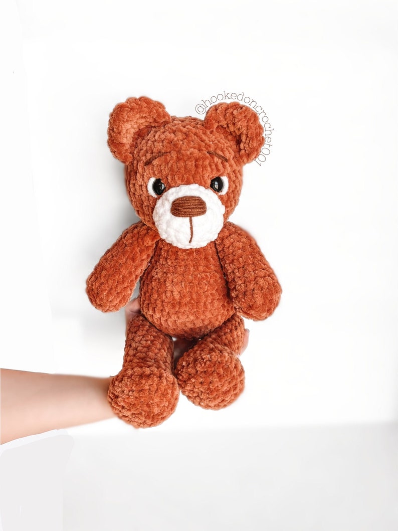 Peut inclure: Un ours en peluche crochet&eacute; marron avec des accents blancs autour des yeux et du nez. L'ours est assis sur une main.
