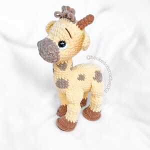 May include: A crocheted giraffe toy with brown spots and a brown nose. The giraffe has large, black eyes and a brown crocheted mane. The toy is standing on a white surface. @hookedoncrochet001