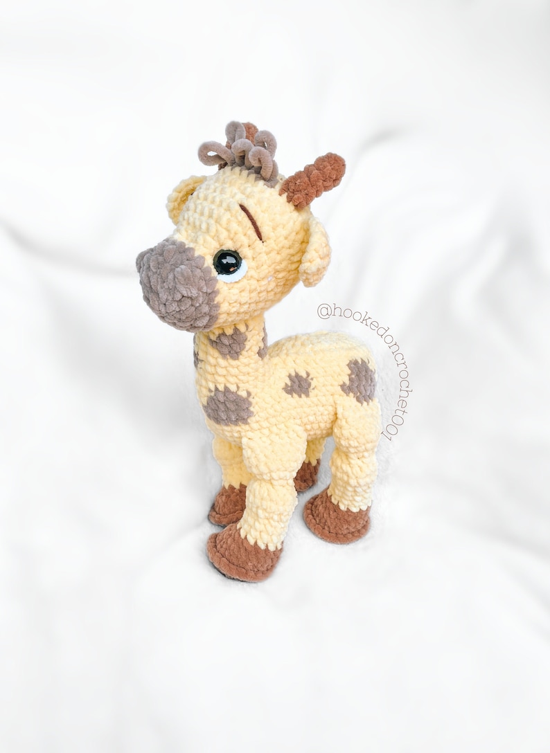 May include: A crocheted giraffe toy with brown spots and a brown nose. The giraffe has large, black eyes and a brown crocheted mane. The toy is standing on a white surface. @hookedoncrochet001