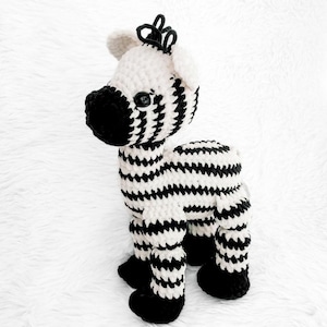 May include: A handmade crochet zebra toy in black and white. The zebra has a black snout, hooves, and a mane. The body is white with black stripes. The toy is standing on a white surface.