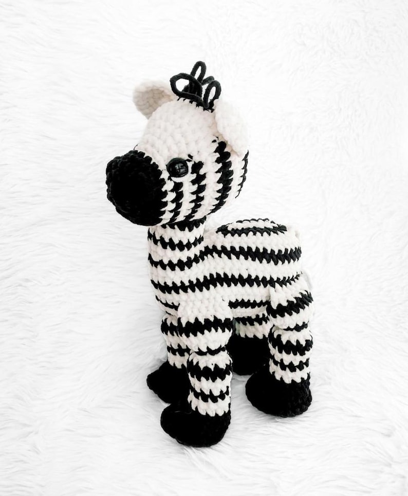 May include: A handmade crochet zebra toy in black and white. The zebra has a black snout, hooves, and a mane. The body is white with black stripes. The toy is standing on a white surface.