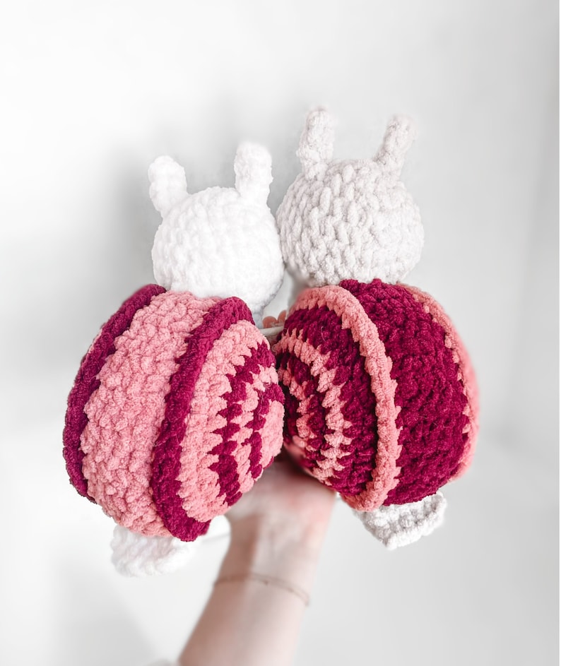 K&ouml;nnte beinhalten: Zwei geh&auml;kelte Schneckenspielzeuge mit rosa und burgunderfarbenen gestreiften Schalen. Die Schnecken haben wei&szlig;e geh&auml;kelte K&ouml;pfe und schauen sich an.