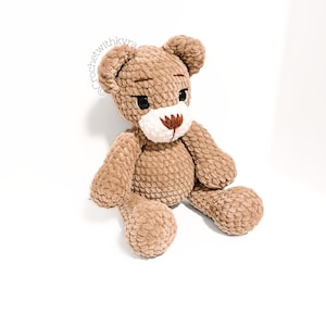 Peut inclure: Un ours en peluche crochet&eacute; marron avec un nez blanc et des yeux brod&eacute;s marrons. L'ours est assis, les jambes tendues.