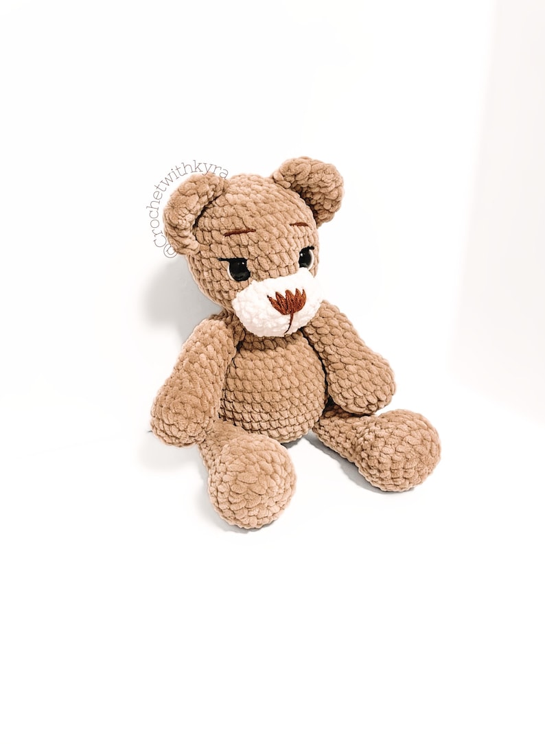 Peut inclure: Un ours en peluche crochet&eacute; marron avec un nez blanc et des yeux brod&eacute;s marrons. L'ours est assis, les jambes tendues.