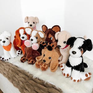 May include: A collection of eight handmade crochet plush dogs in various colors including white, brown, black, and tan. Each stuffed animal features soft yarn, dark plastic eyes, and distinct ear shapes, arranged together in a group on a white surface.