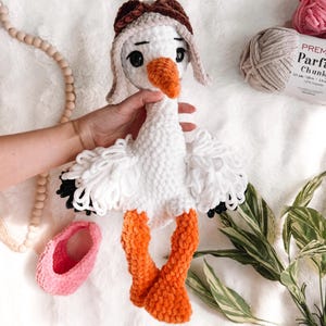 Puede incluir: Un peluche de cigüeña tejido a ganchillo en color blanco con cuello largo, pico naranja y patas naranjas. La cigüeña lleva un gorro de aviador marrón con orejeras. Las alas están hechas de bucles de hilo blanco con puntas negras y ojos negros.