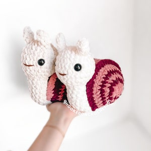 K&ouml;nnte beinhalten: Zwei geh&auml;kelte Schnecken, eine wei&szlig; und eine mit einer rosa-burgunderfarbenen Schale. Die Schnecken halten sich aneinander fest und werden von einer Hand hochgehalten. Der Text "@hooked.on.crochet.001" ist in der rechten unteren Ecke des Bildes sichtbar.