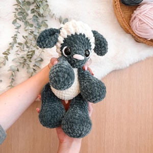 May include: A handmade crochet sheep plushie in shades of gray and white. The sheep has large, round eyes and a pink nose. The plushie is held in someone's hands. The sheep is a soft, cuddly toy.