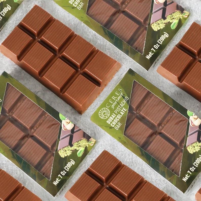 May include: Several milk chocolate bars are arranged on a light surface. Each bar is segmented into rectangular pieces. The packaging includes the text "Dubai Pistachio Chocolate Bar" and "Net: 7 Oz (200g)".